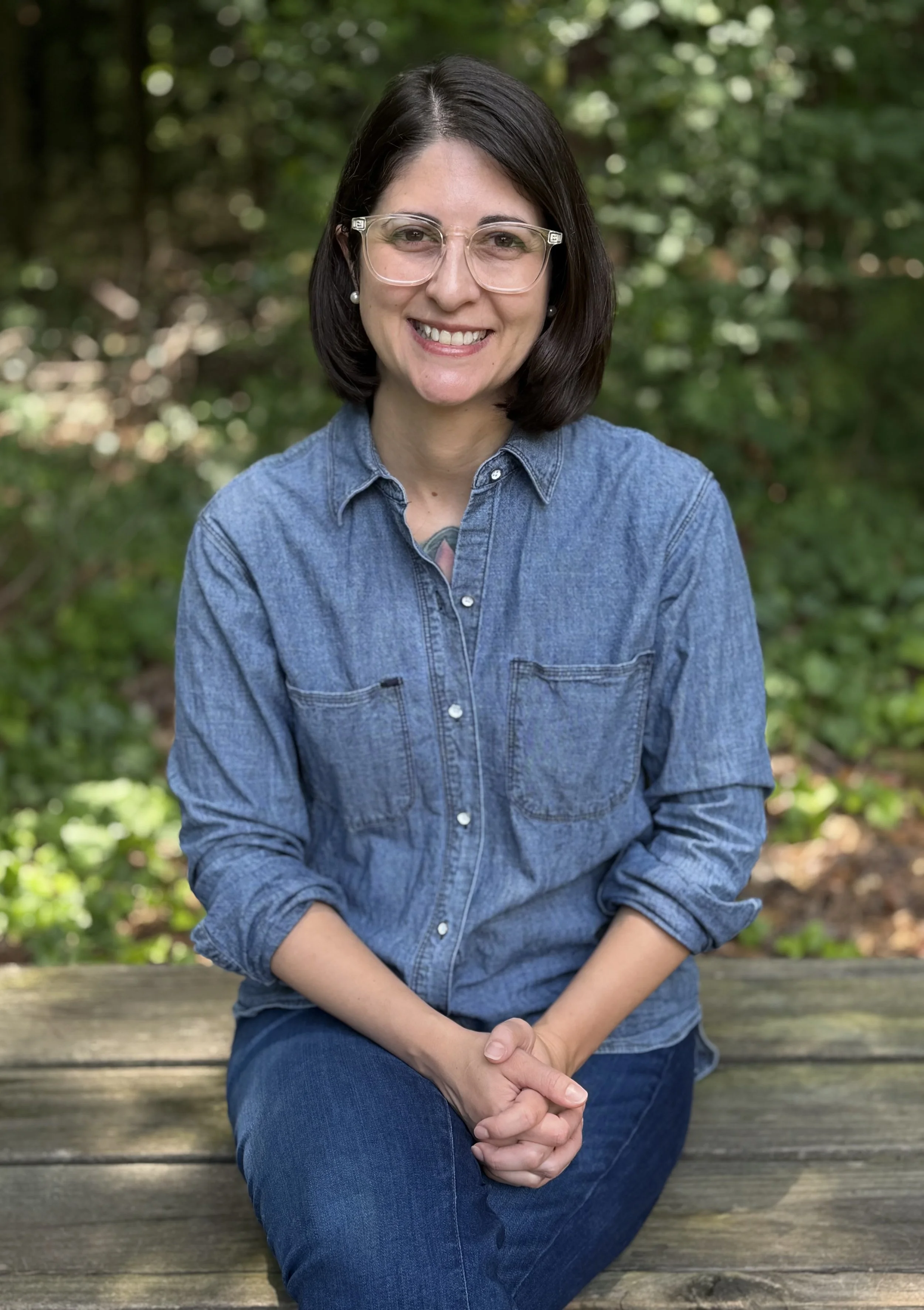 A woman with shoulder-length dark hair, wearing glasses, pearl earrings, a denim shirt, and jeans, sitting outdoors on a wooden bench with green foliage background, smiling at the camera.