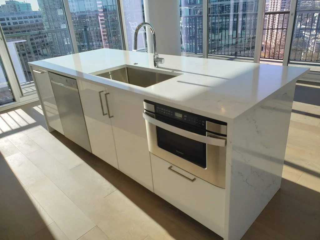 Modern kitchen island with white marble countertop, built-in sink, stainless steel dishwasher, and oven, in a high-rise apartment with large windows and city views.