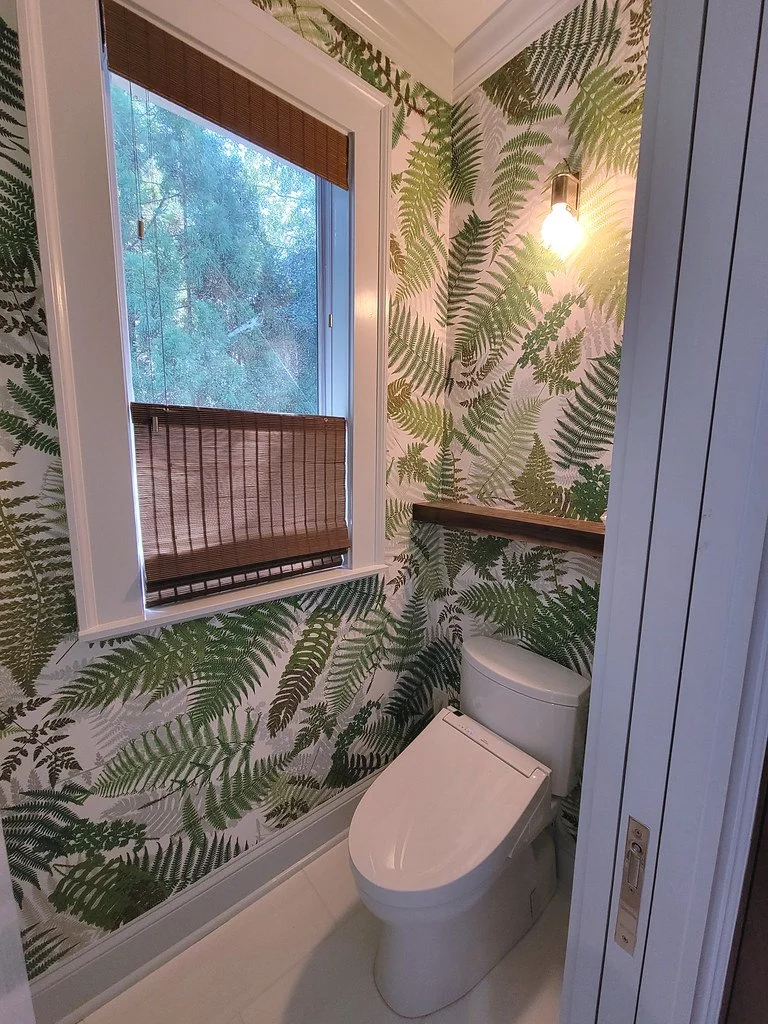 A small bathroom with a wall covered in fern-patterned wallpaper, a toilet, a window with bamboo blinds, a wooden shelf near the toilet, and a wall-mounted light fixture.