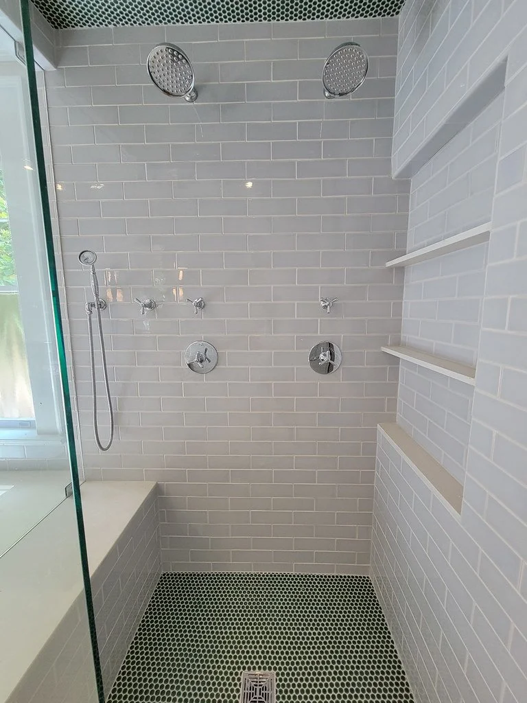 Modern walk-in shower with gray subway tile walls, two large overhead shower heads, a handheld shower head, and built-in shelves. The floor has small round black tiles with a drain in the center.