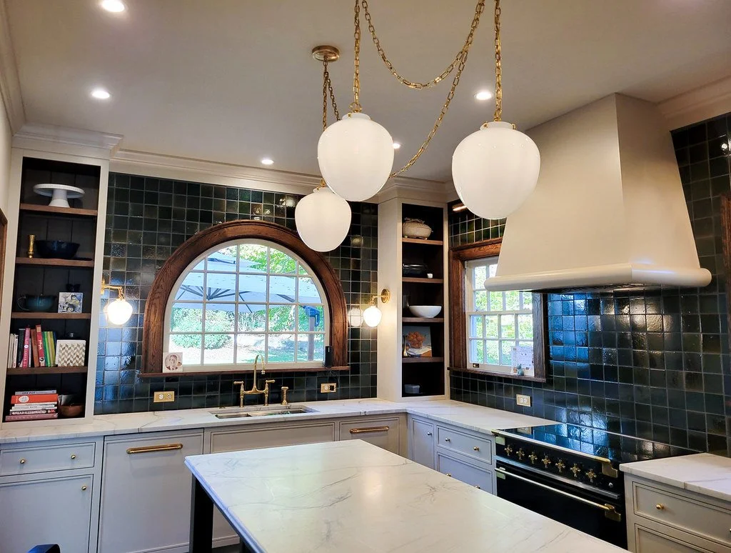 Kitchen with dark green tiled backsplash, white cabinets with gold hardware, a marble countertop island, brass chandelier with three globe lights, arched window with a view of greenery, and a white range hood over a black and gold oven.