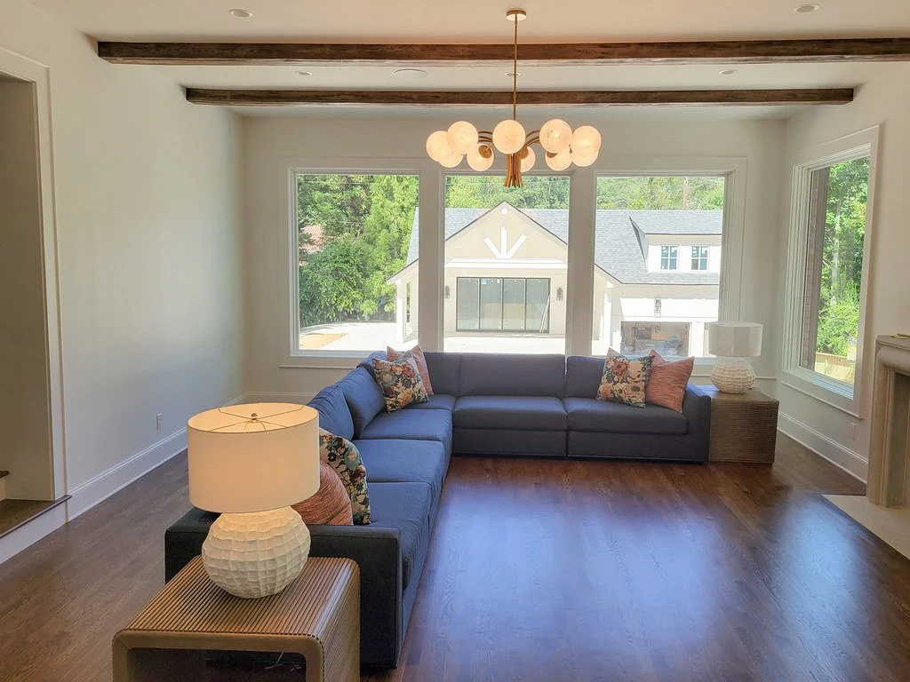Living room with a navy sectional sofa, colorful throw pillows, two table lamps, large windows showing a view of neighboring houses and greenery, hardwood floor, and a modern chandelier hanging from the ceiling.