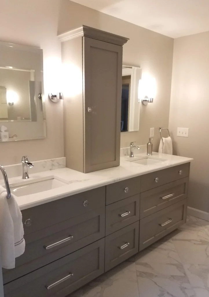 Double bathroom vanity with grey cabinets, white marble countertop, two sinks, and two mirrors. There are wall-mounted light fixtures and a tall grey cabinet between the mirrors. The bathroom has tiled flooring and neutral-colored walls.