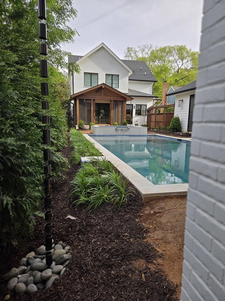 Backyard with a swimming pool, a wooden pool house, and a white two-story house in the background. There are landscaped bushes and plants along the edge of the yard.