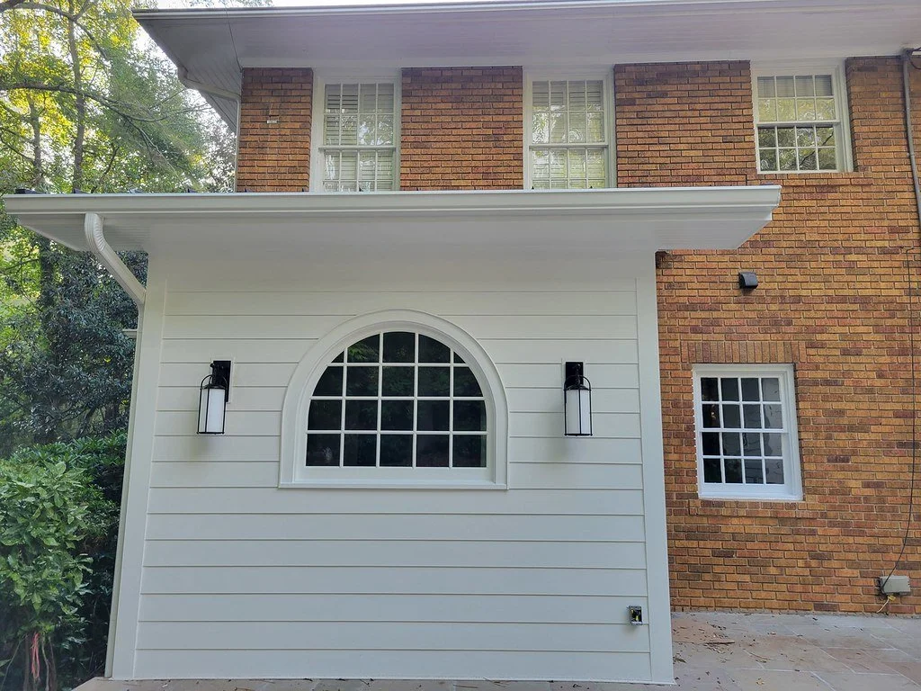 Back of a brick house with a white section featuring a large arched window and two black outdoor wall lanterns, set against a background of green trees.