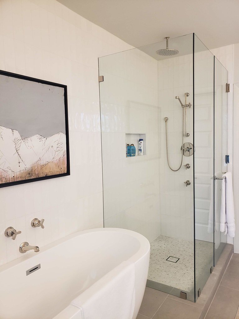 A modern bathroom with a white bathtub, a glass-shower enclosure with a rain showerhead, built-in shower niche with toiletries, towel rack with a white towel, and a framed black and white landscape art on the wall.