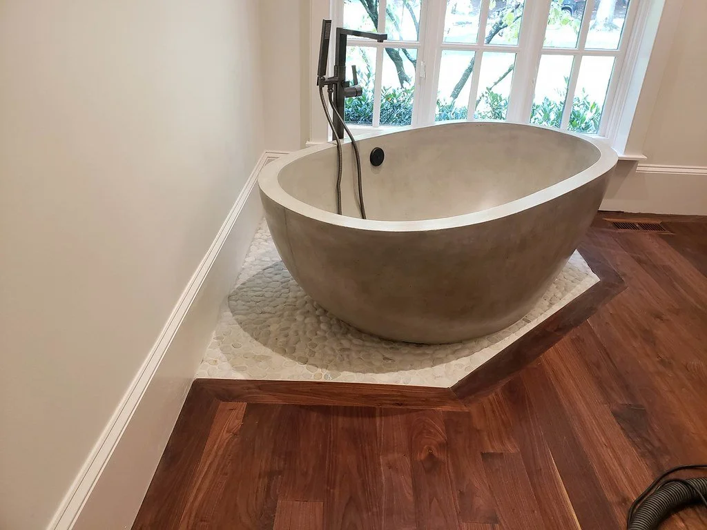 A modern bathroom with a freestanding oval bath, a large window with a garden view, and hardwood flooring.