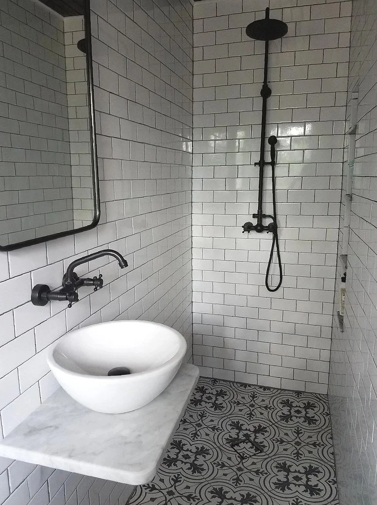 A bathroom with white subway tile walls, a round white vessel sink on a gray marble countertop, wall-mounted black faucet, a large mirror, and a separate walk-in shower area with a black overhead shower head and hand-held shower attached to the wall,