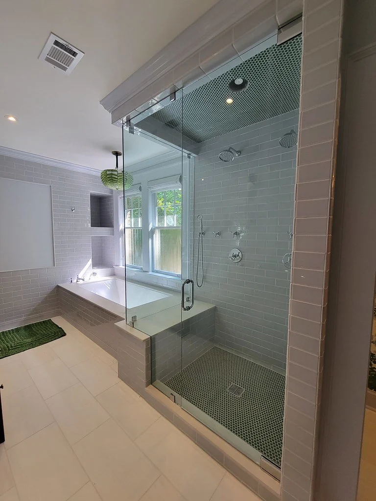 Modern bathroom with glass-enclosed shower featuring two showerheads and a built-in bathtub next to a window with white frames, white tiled walls, and a light-colored tiled floor, with a green bath mat on the floor.