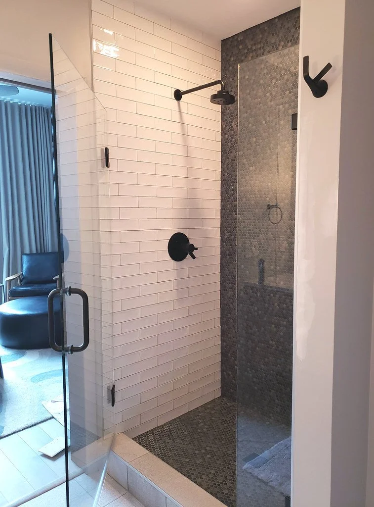 Modern walk-in shower with white subway tiles on the left wall and dark pebble tiles on the floor and back wall. The shower has a glass door and black fixtures, including a showerhead and control knob.