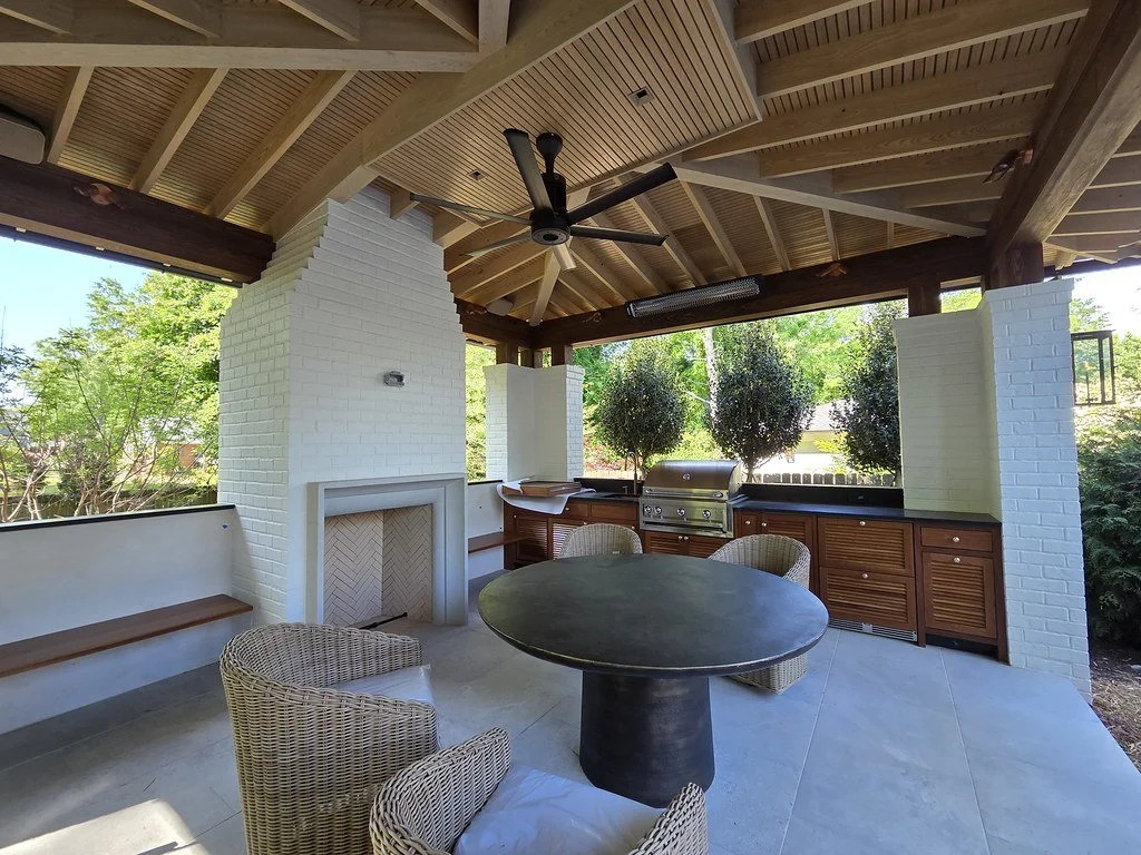 Outdoor covered patio with a black round table, wicker chairs, built-in bench, white brick fireplace, ceiling fan, outdoor grill, and surrounding trees.