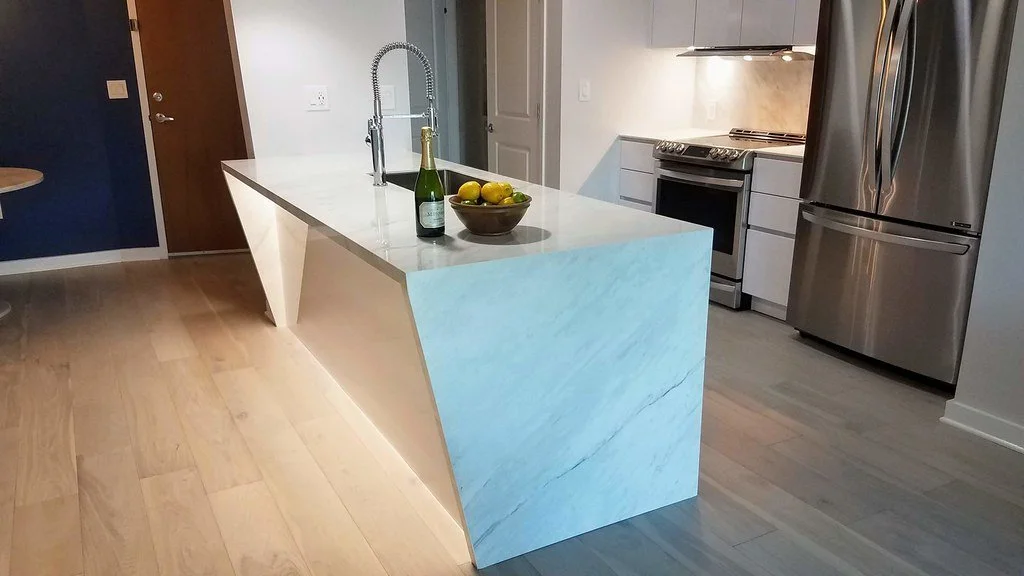Modern kitchen with white marble island, stainless steel refrigerator, oven, and light-colored wood flooring.