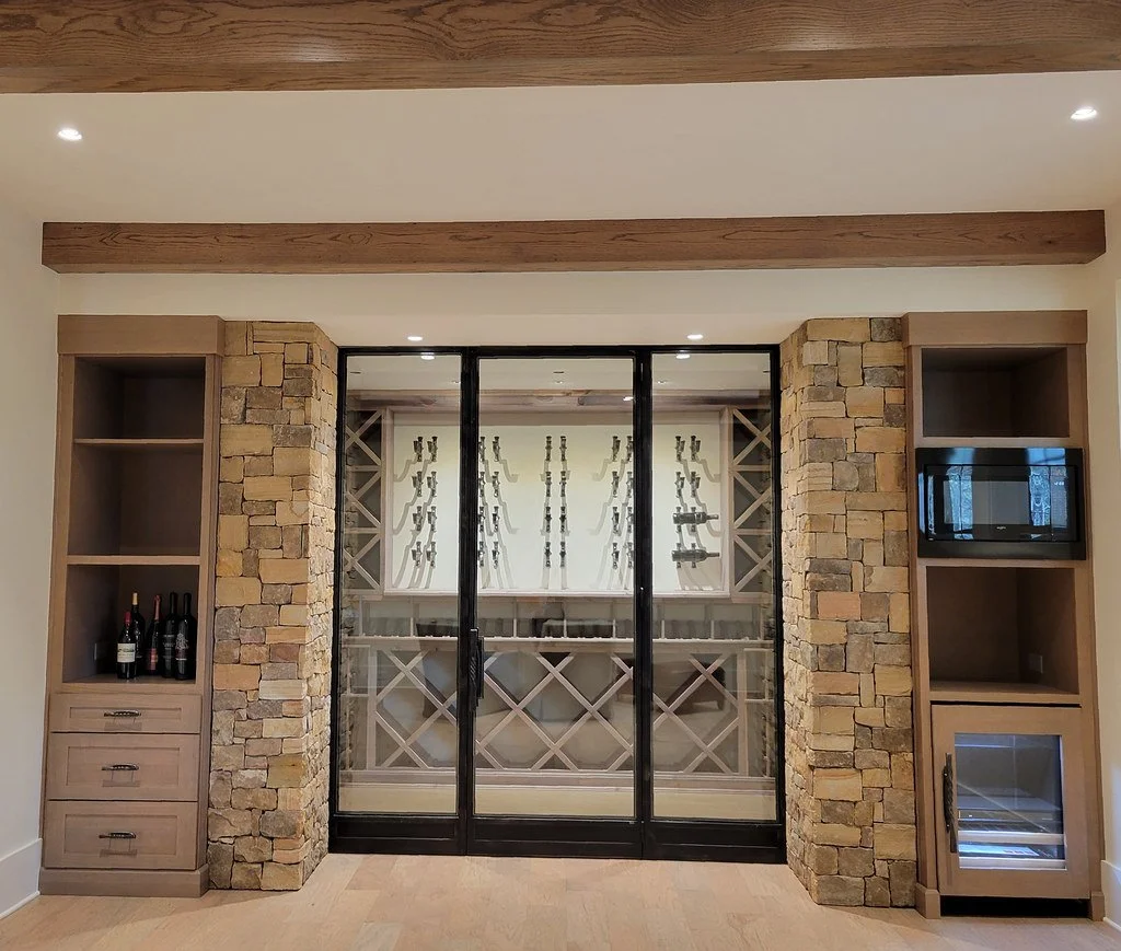 Living room with stone and wood accents, glass sliding doors leading to a wine cellar, built-in shelves with wine bottles and a mini-fridge, and ceiling with recessed lighting.