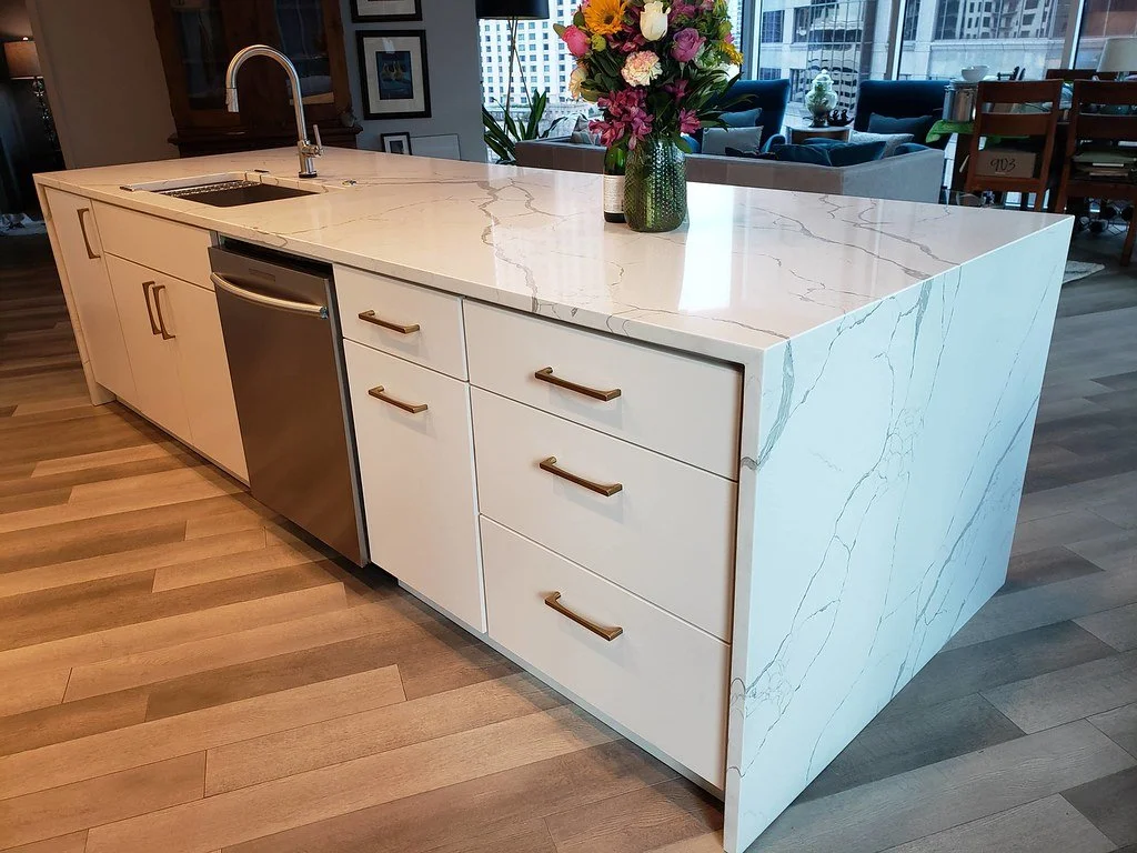 Modern kitchen island with white marble countertop, drawers with gold handles, a small sink, and a vase with a colorful flower arrangement. In the background, there are dining and living areas with large windows.