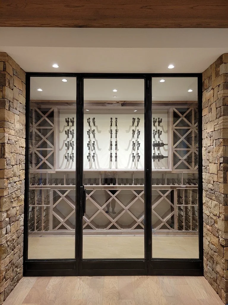 View of a glass wine cellar door with black framing, showing a collection of firearms displayed on the back wall, flanked by stone walls on either side.