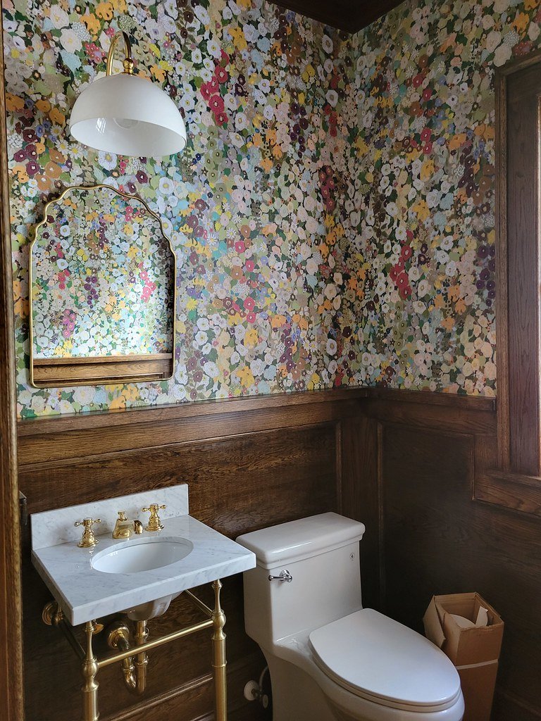 A small bathroom with floral wallpaper, a wall-mounted mirror with a gold frame, a white marble sink with gold fixtures, a white toilet, a window, and a small trash bin.