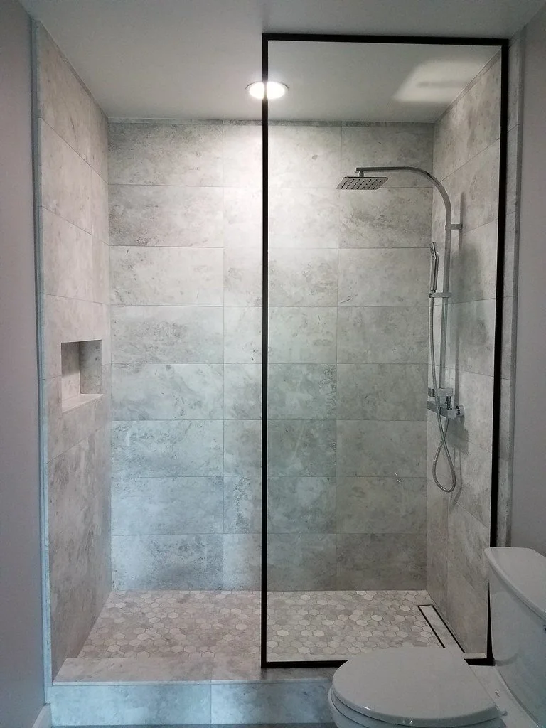 Modern walk-in shower with gray tile walls, a built-in niche, and a rain shower head, separated by a glass partition, with a white toilet nearby.
