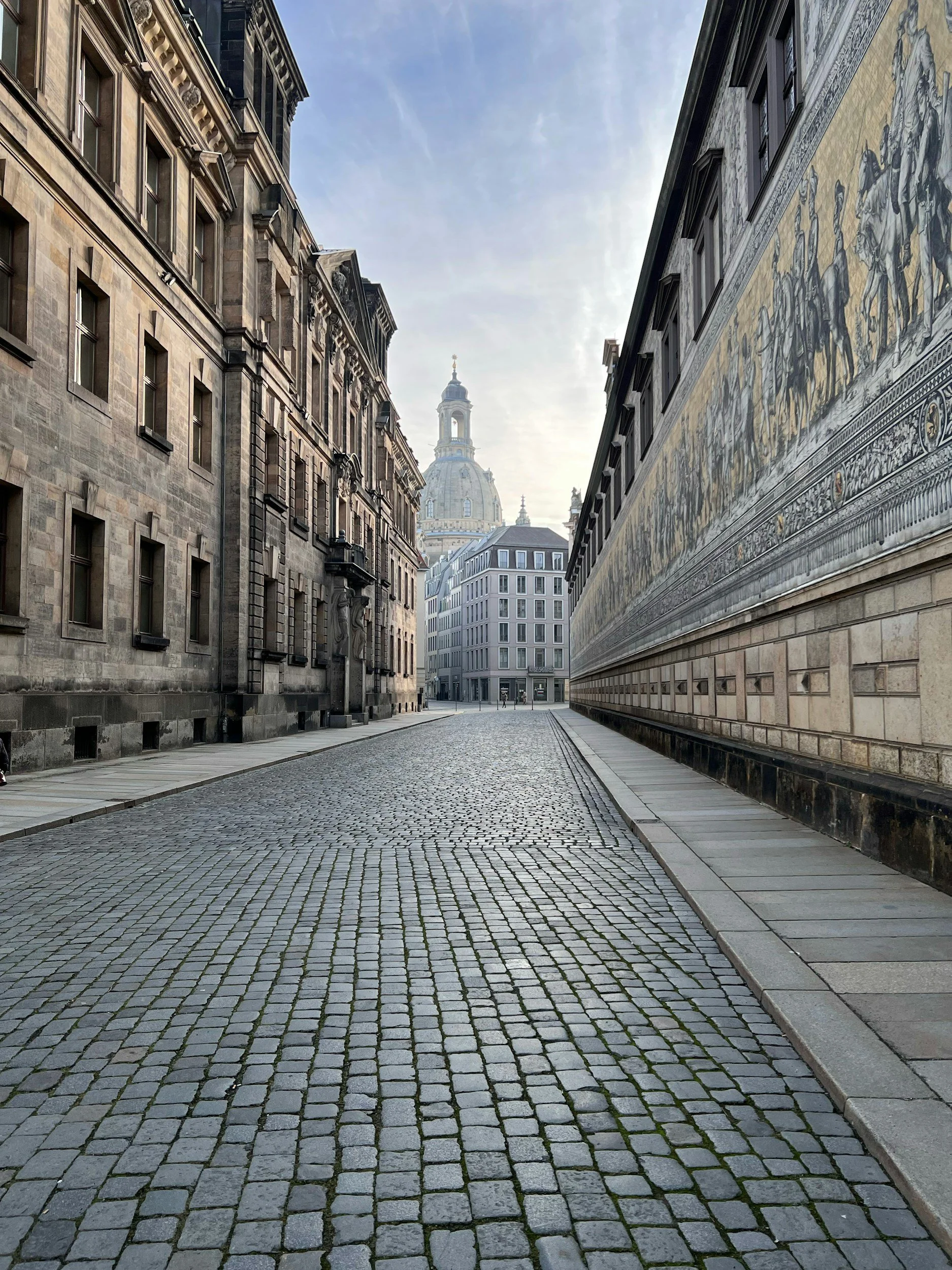 Empty cobblestone street flanked by historic buildings, with a domed church visible in the background under a partly cloudy sky. Location in Dresden, home of Sachsenküchen. Premium German kitchens.