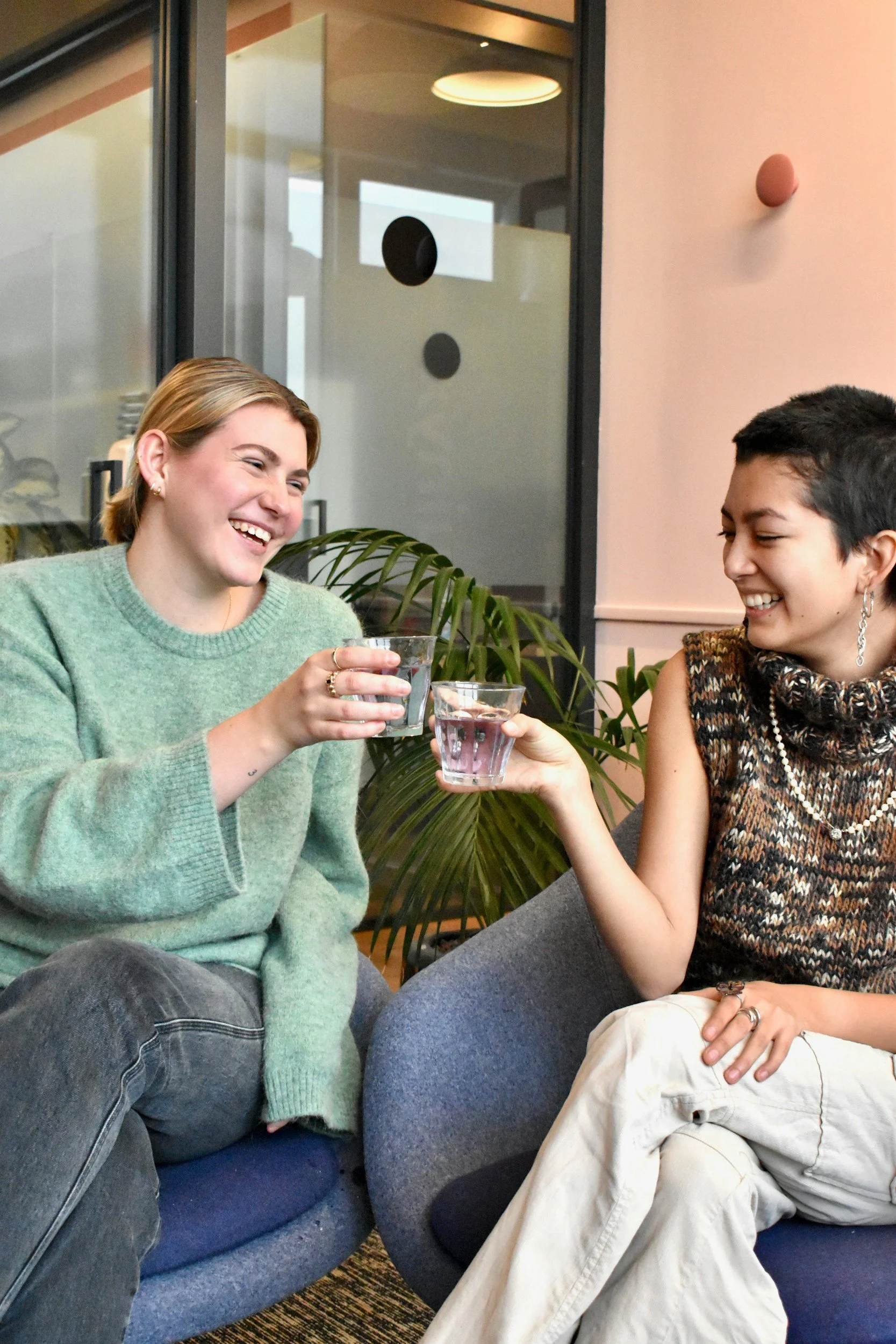 Two women sitting on a blue sofa, smiling and holding glasses of water, toasting. The woman on the left has short blonde hair, is wearing a green sweater, and has earrings. The woman on the right has short dark hair, is wearing a patterned sleeveless top and white pants with holes. They are in a modern indoor space with a glass partition, plants, and soft lighting.
