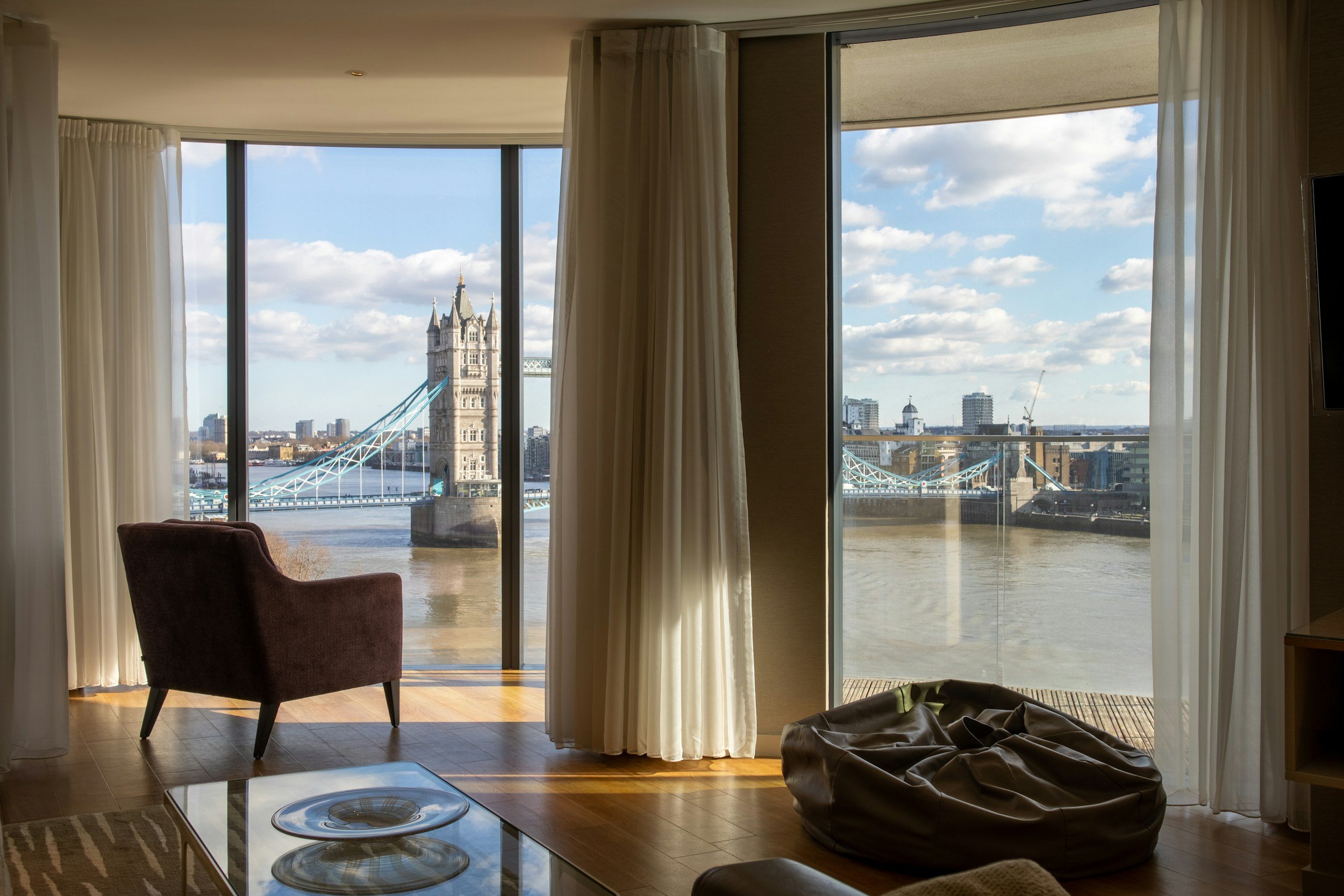View from a hotel room with large windows showing the Tower Bridge in London, with a chair and a floor lamp inside the room, and a crumpled blanket on the floor. Luxury homes and apartments. Premium luxury furniture.