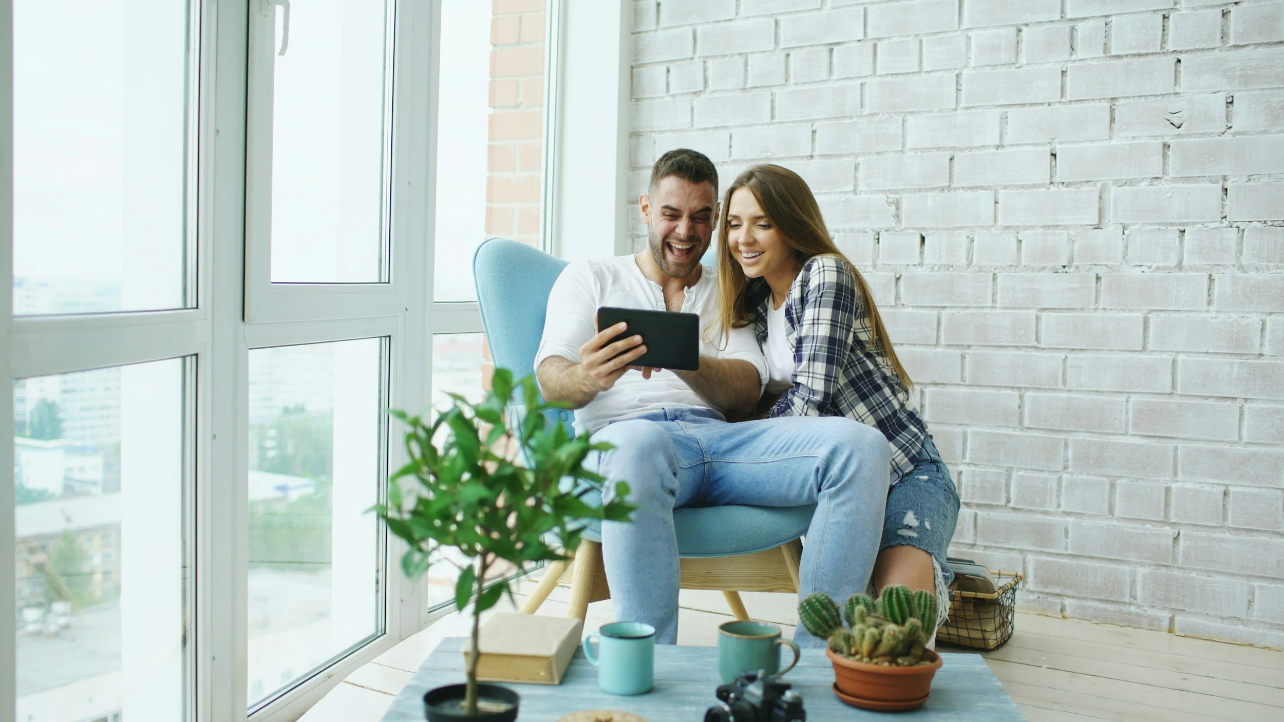 A young man and woman sitting on a chair by a large window, looking at a tablet and laughing together.