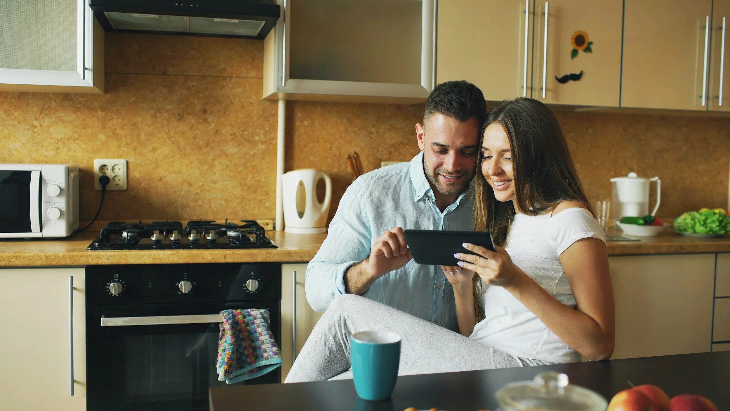 A man and woman sitting together in a kitchen, looking at a smartphone. The kitchen has beige cabinets, a stove, a microwave, and a counter with kitchen items.