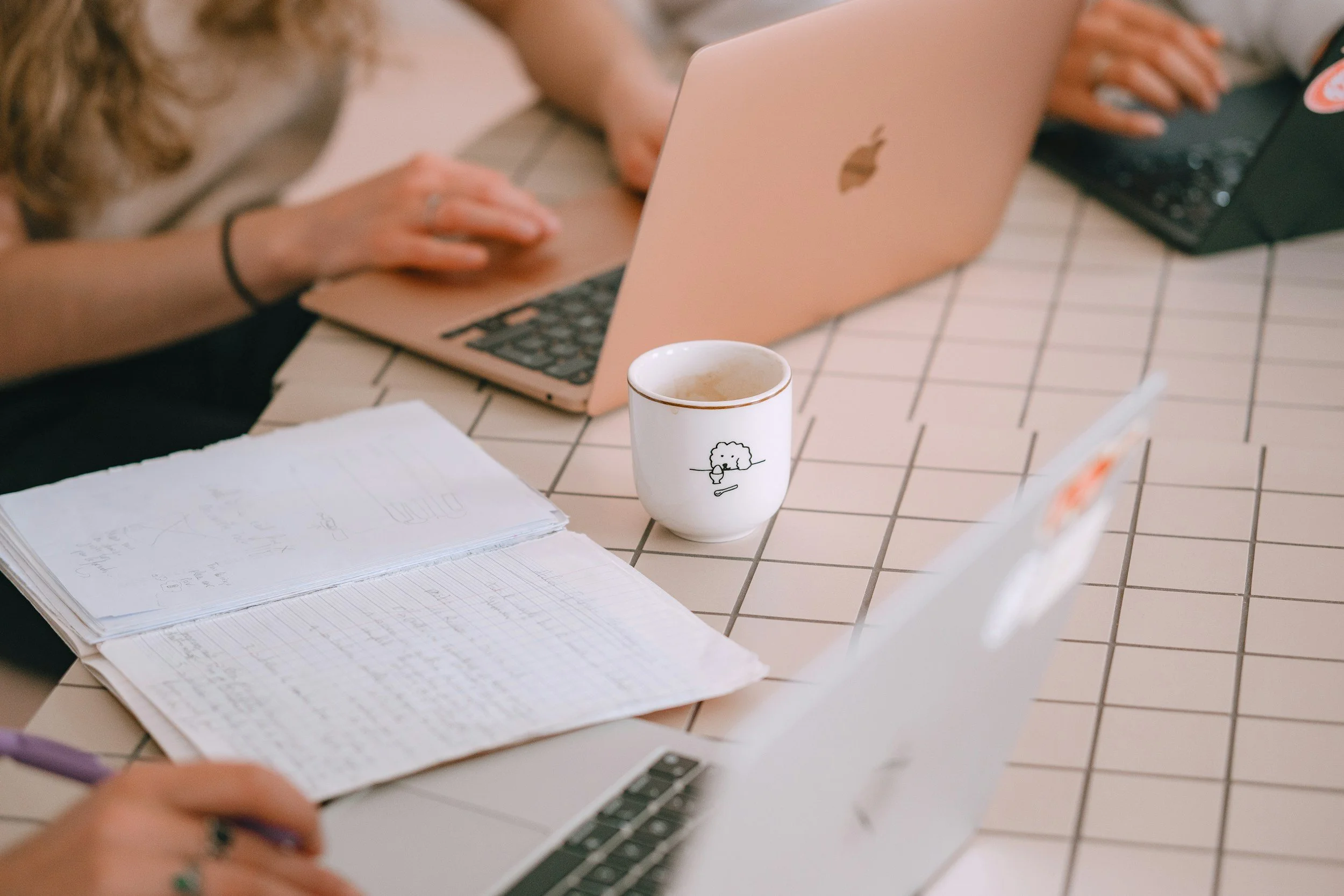 Close-up of a workspace on a tiled table with a notebook, a pen, a mug with a sheep illustration, a person typing on a pink laptop, and another person reaching for a black device.