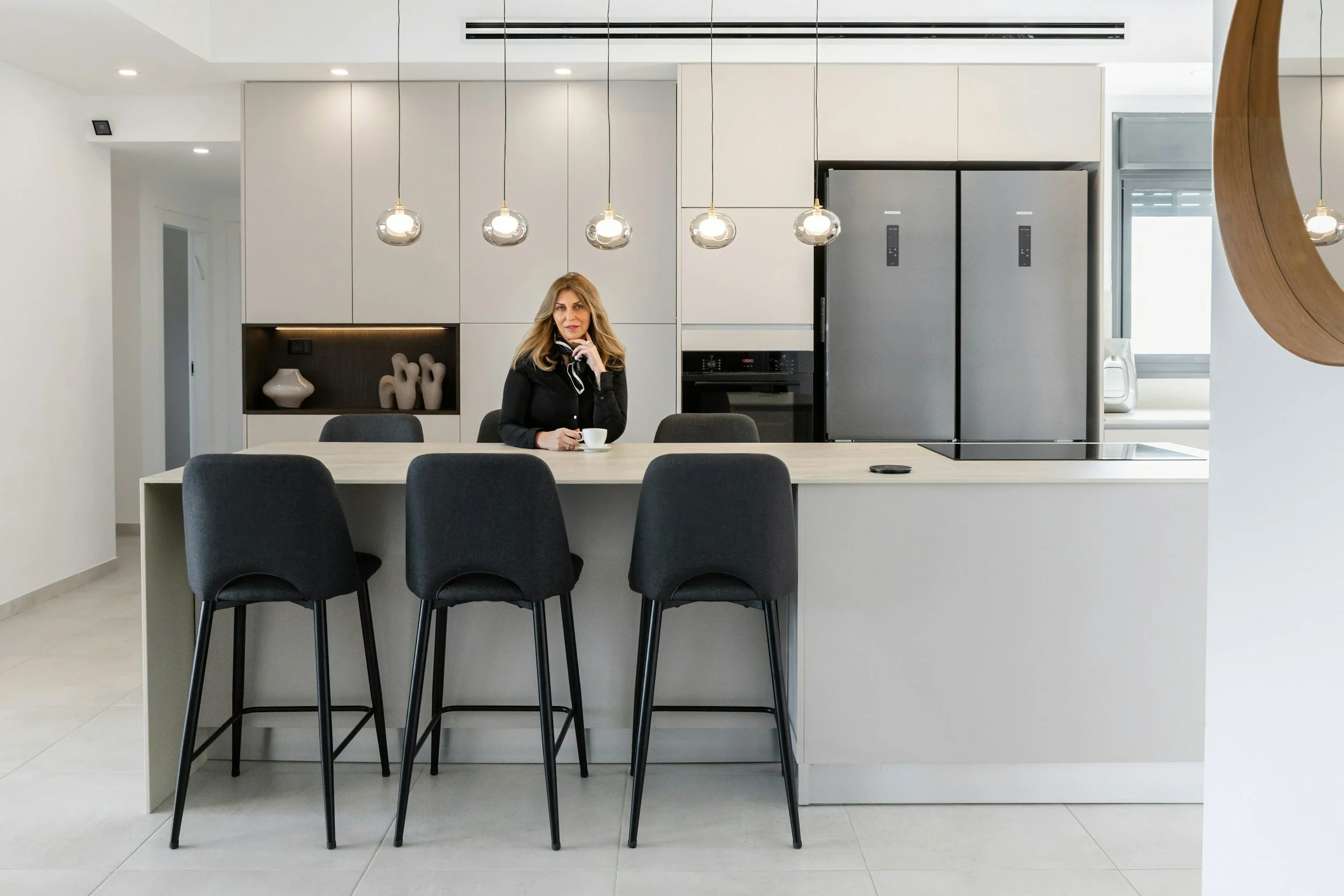 Modern kitchen with a woman sitting at the counter, white cabinets, stainless steel appliances, hanging pendant lights, and black bar stools.