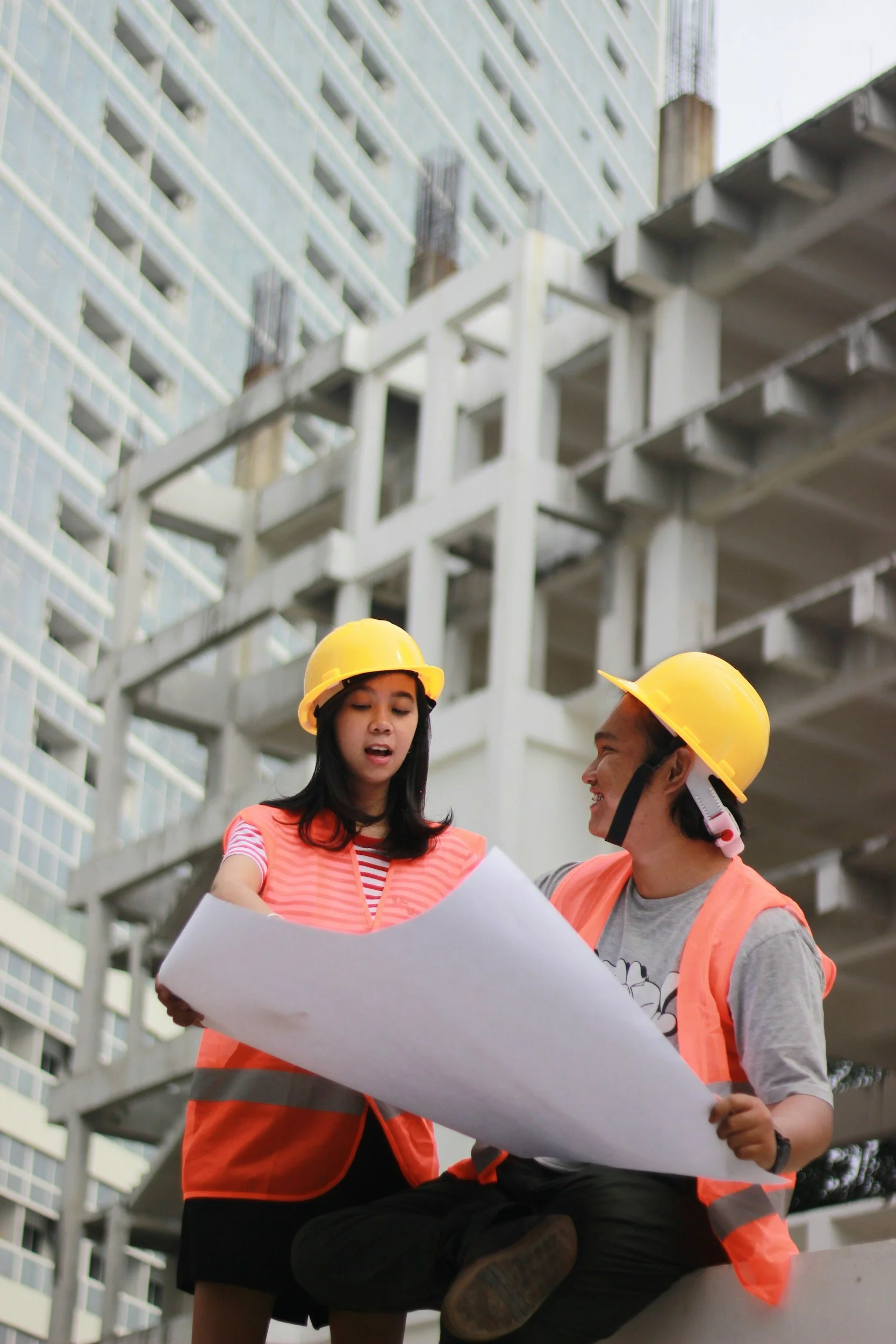 Two construction workers, a woman and a man, wearing yellow safety helmets and orange safety vests, reviewing blueprints at a construction site with a building under construction in the background.