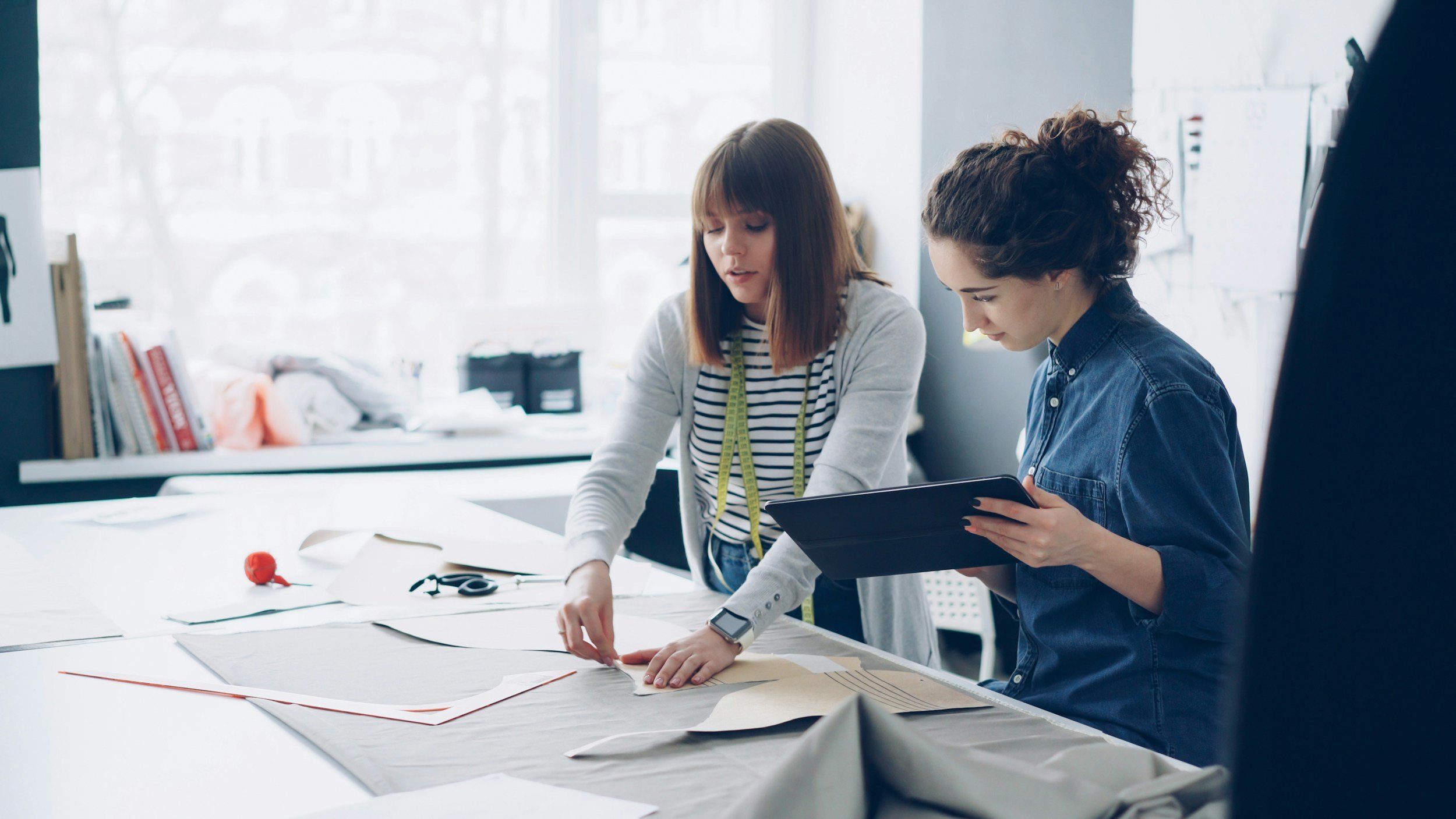Two women work together on a sewing or pattern project at a large table in a bright, modern workspace. One woman in a striped shirt with a measuring tape around her neck is pointing at a pattern or fabric, while the other woman in a denim shirt holds a tablet.