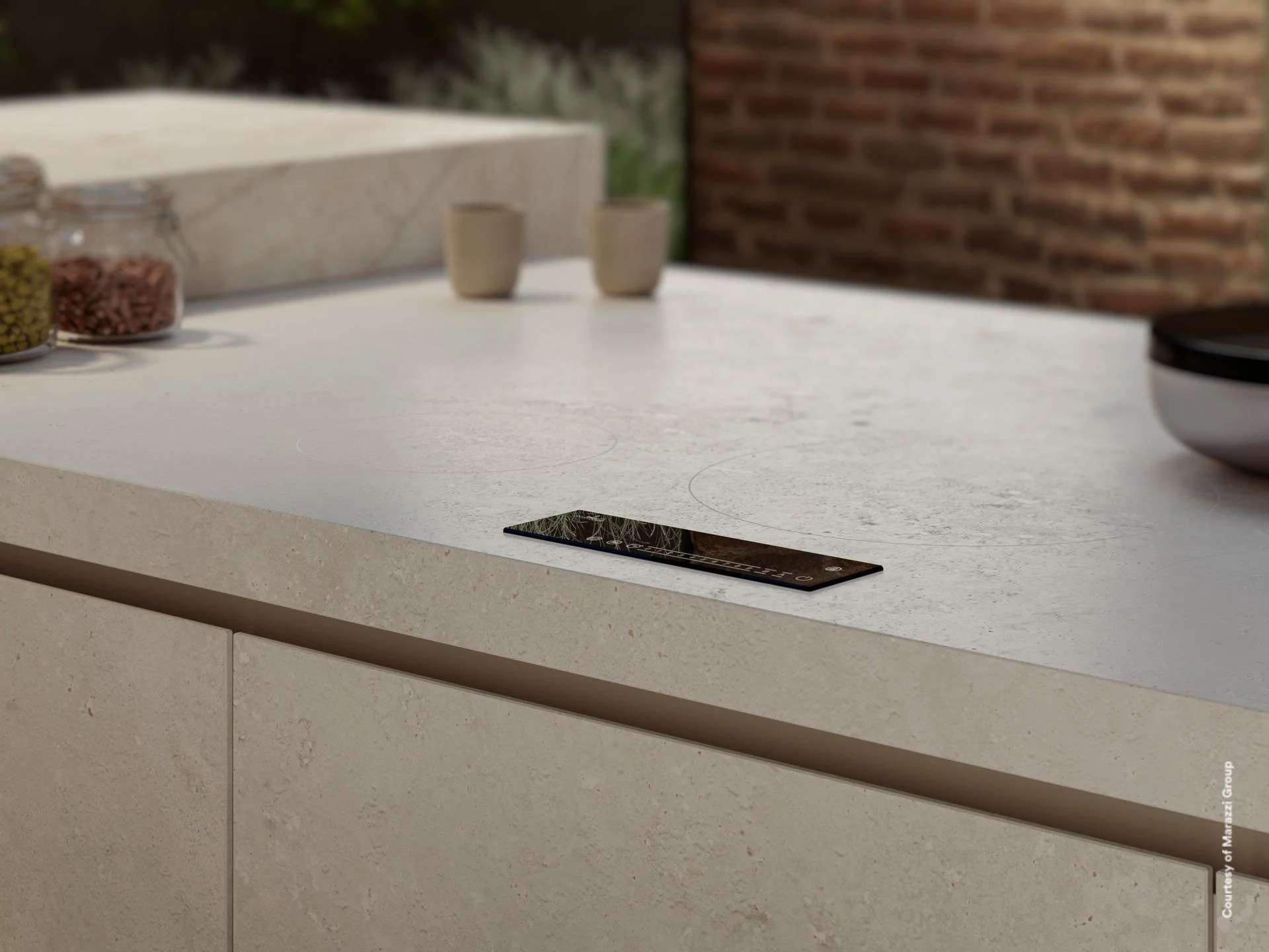 Modern kitchen countertop with a black induction stove, two beige cups, jars of spices, and a white textured surface, with a brick wall and greenery in the background.
