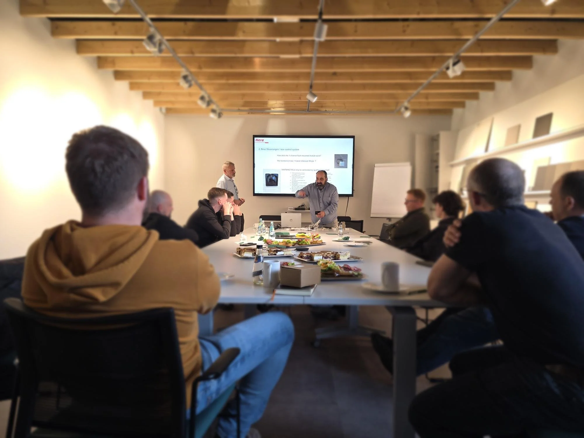 A group of people attending a presentation in a conference room with a wooden ceiling, where a man is standing in front of a large screen displaying a slide, and others are sitting at a table with food and drinks.
