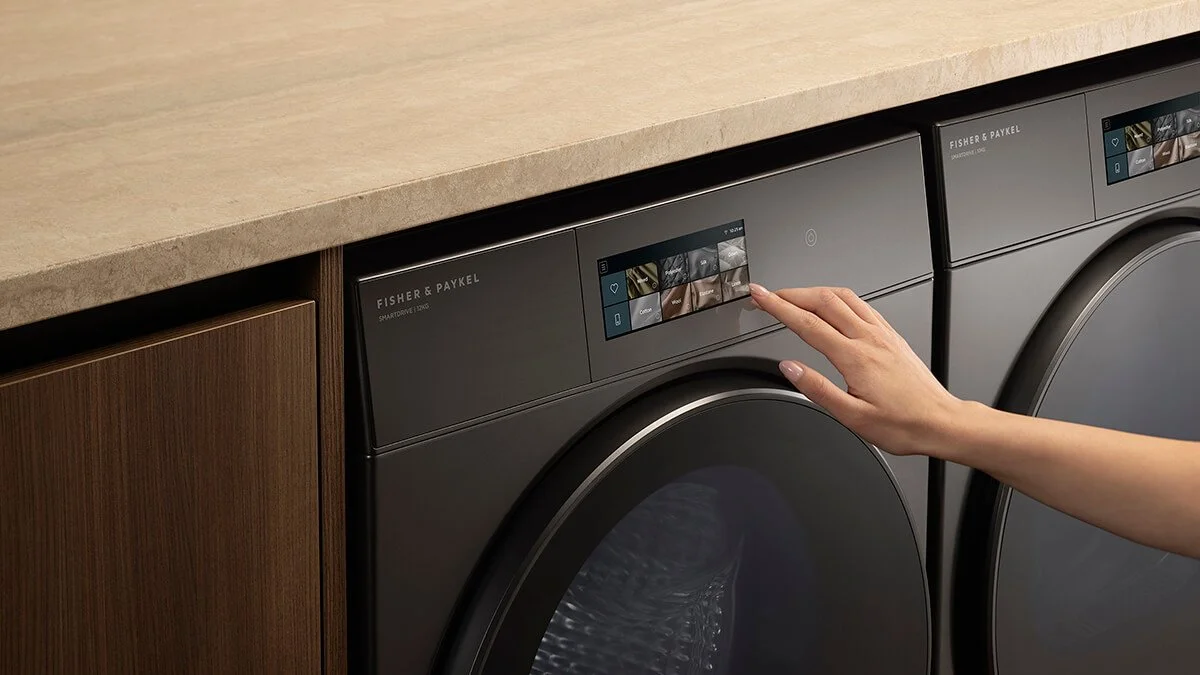 A person pressing a touchscreen control panel on a black Fisher & Paykel front-load washing machine, located beside a matching dryer, in a laundry room with a beige countertop and wooden cabinetry.