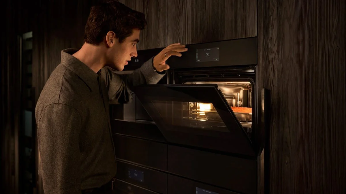 A man opening and inspecting an oven with a tray of food inside, located in a modern kitchen with dark wood paneling.