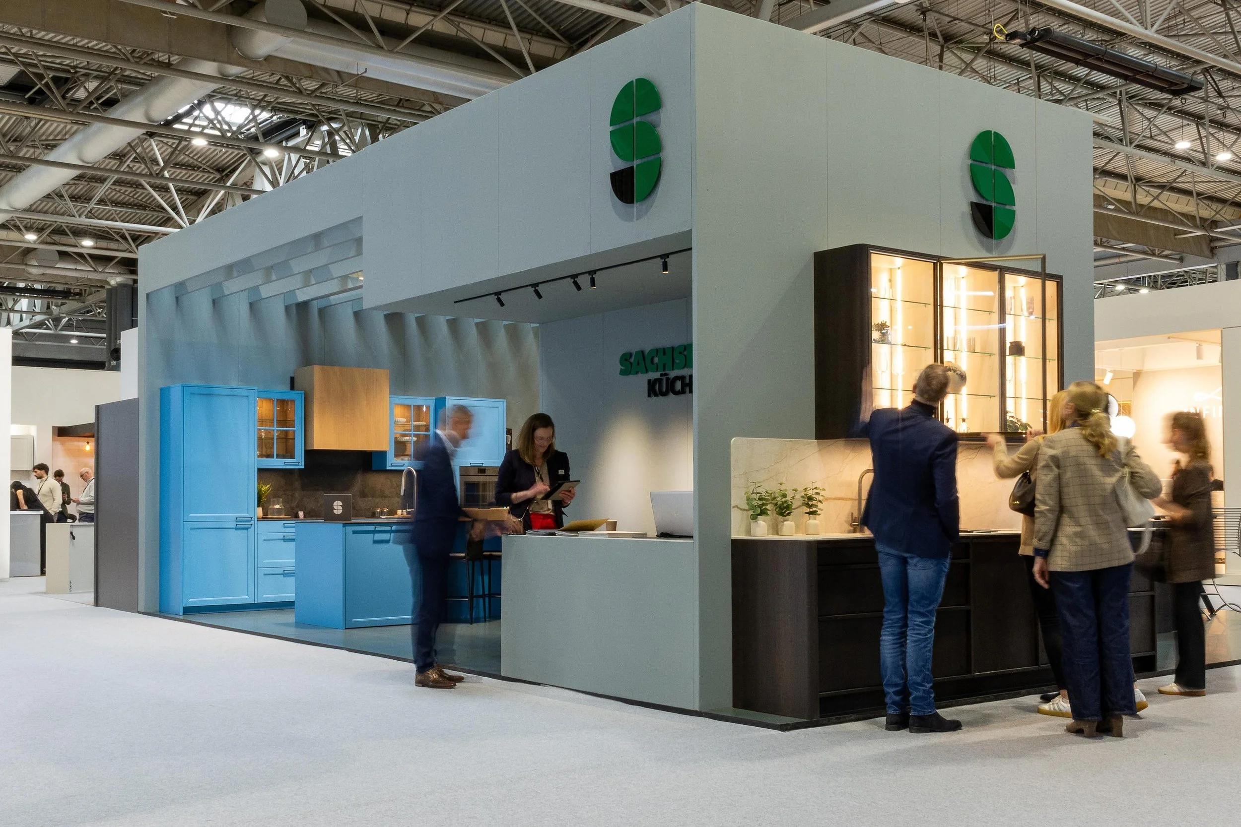Trade show booth with blue kitchen cabinets and a gray display structure, featuring the logo of a green and black circle. Several people are talking and walking around in the booth area in a large exhibition hall with a high ceiling and visible ventilation ducts.