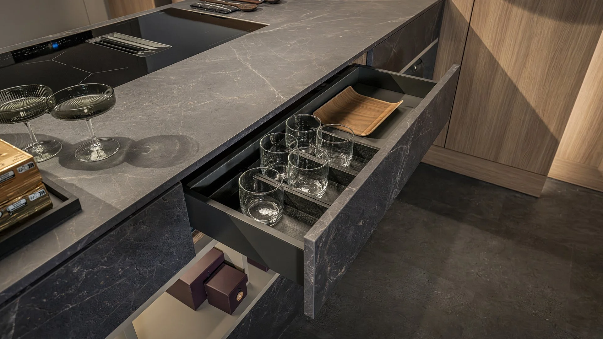 Open kitchen drawer with six empty drinking glasses and a wooden tray inside, adjacent to a marble countertop with glasses and a black ceramic cooktop.