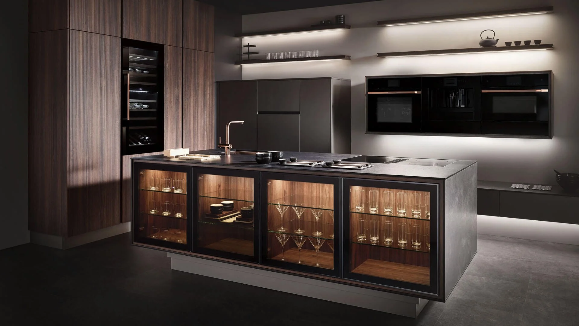 Modern kitchen with dark wood cabinetry, black appliances, a sleek island with glass-front cabinets displaying glassware, and open shelving with kitchenware, illuminated by ambient lighting. ADINA / RITA / FIORA. Sachsenküchen premium kitchens.