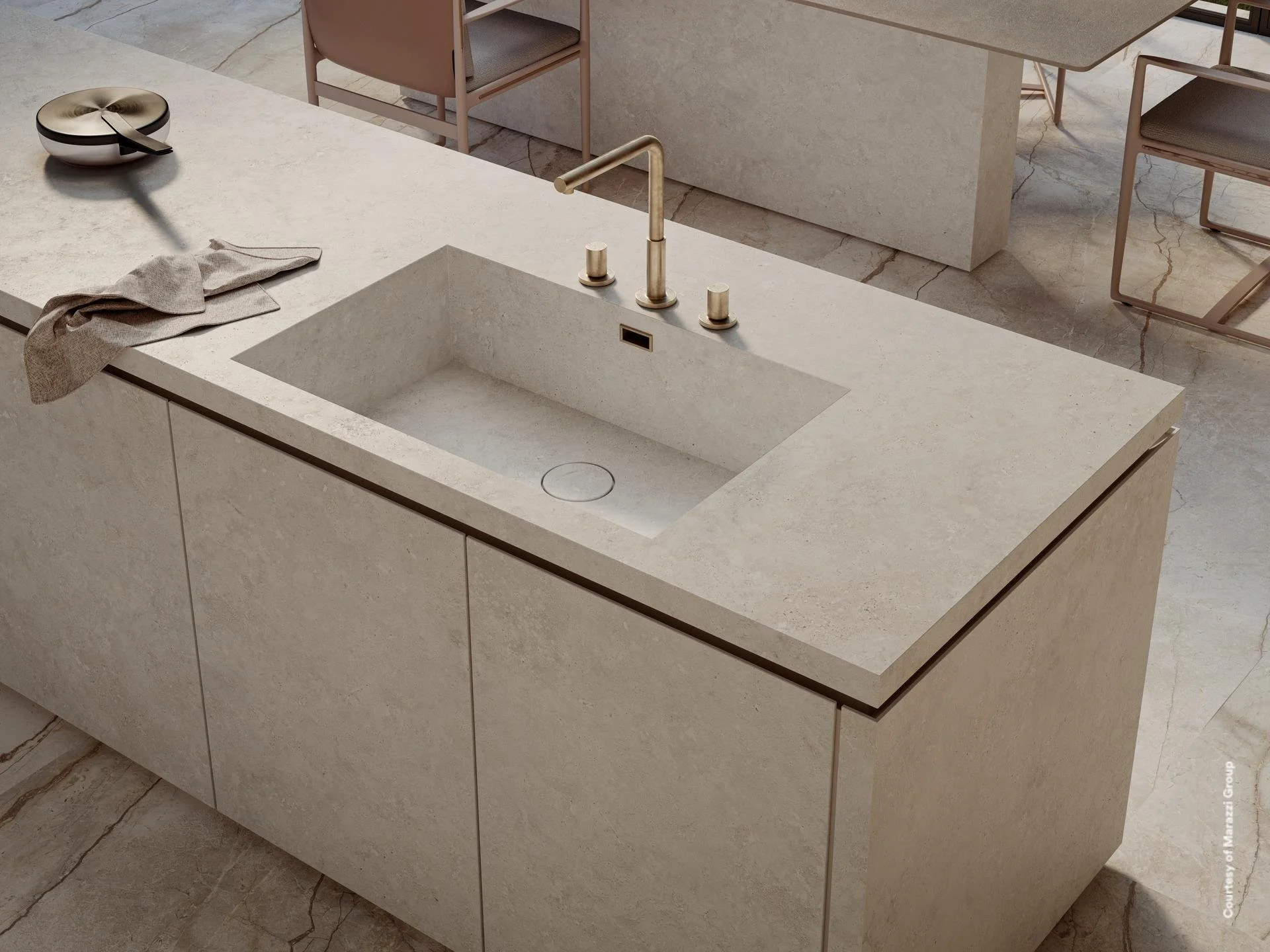 A modern kitchen island with a built-in sink, a gold-coloured faucet, and beige tile surfaces. There are pink chairs and a table in the background. Marazzi premium work surfaces. Luxury worktops. Bespoke German Kitchens.