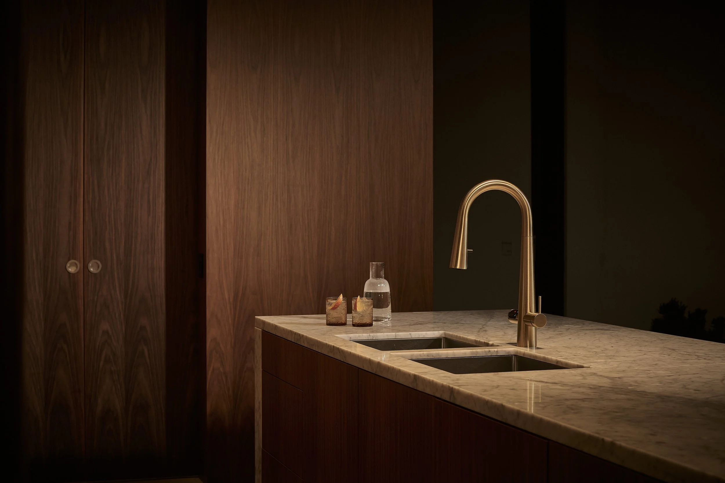 View of a modern kitchen countertop with two glasses of iced drinks, a water bottle, a brass faucet, and wooden cabinets in the background at night.