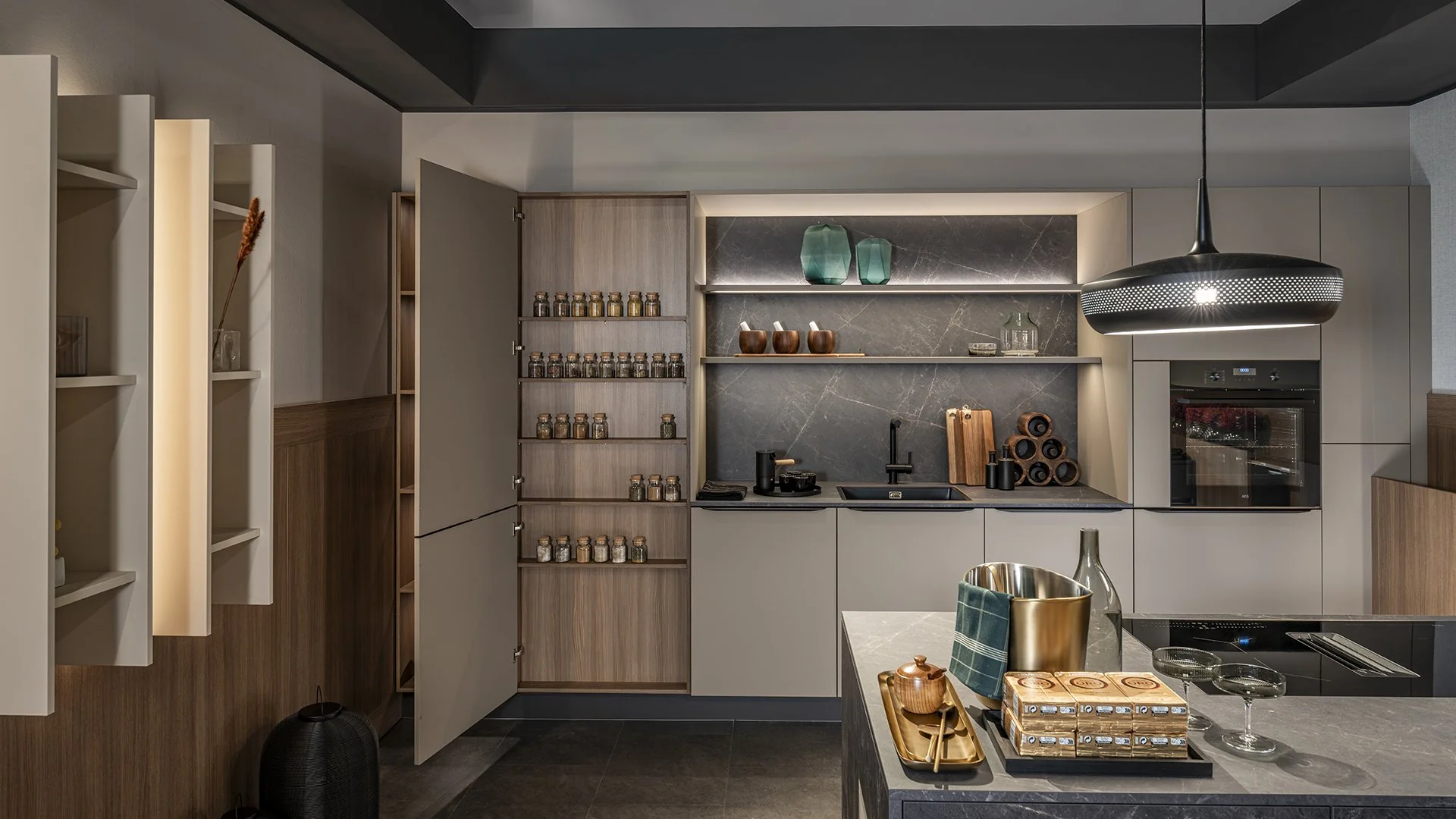 Modern kitchen with built-in oven, open shelving with decorative items, gray and black color scheme, and a kitchen island with tableware
