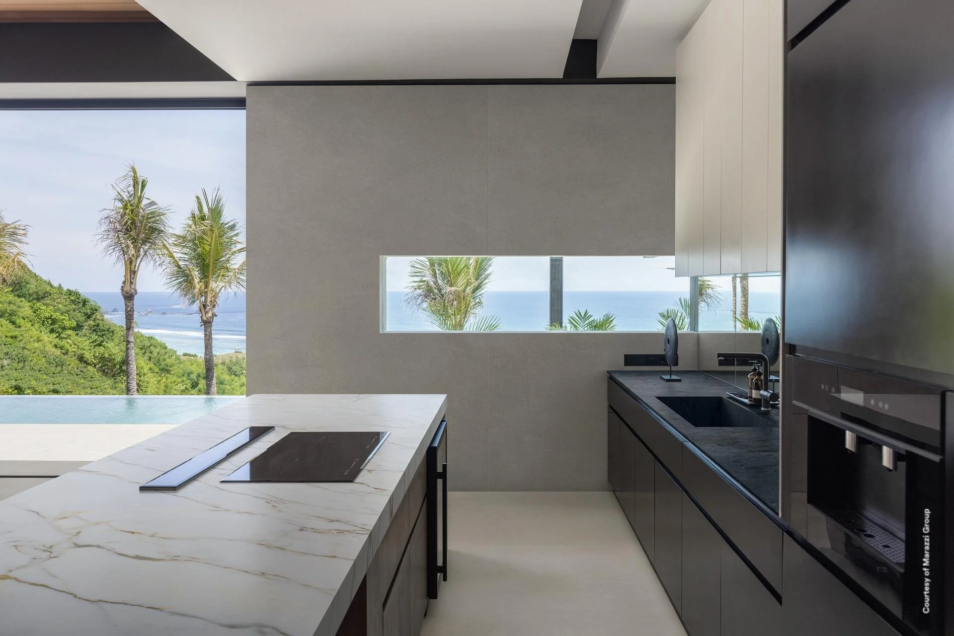 Modern kitchen with a white marble countertop, black induction stove, and large window showing a view of palm trees, greenery, the ocean, and a clear sky. Premium worktops by Marazzi. Best worksurfaces. Sachsenküchen bespoke luxury kitchens.