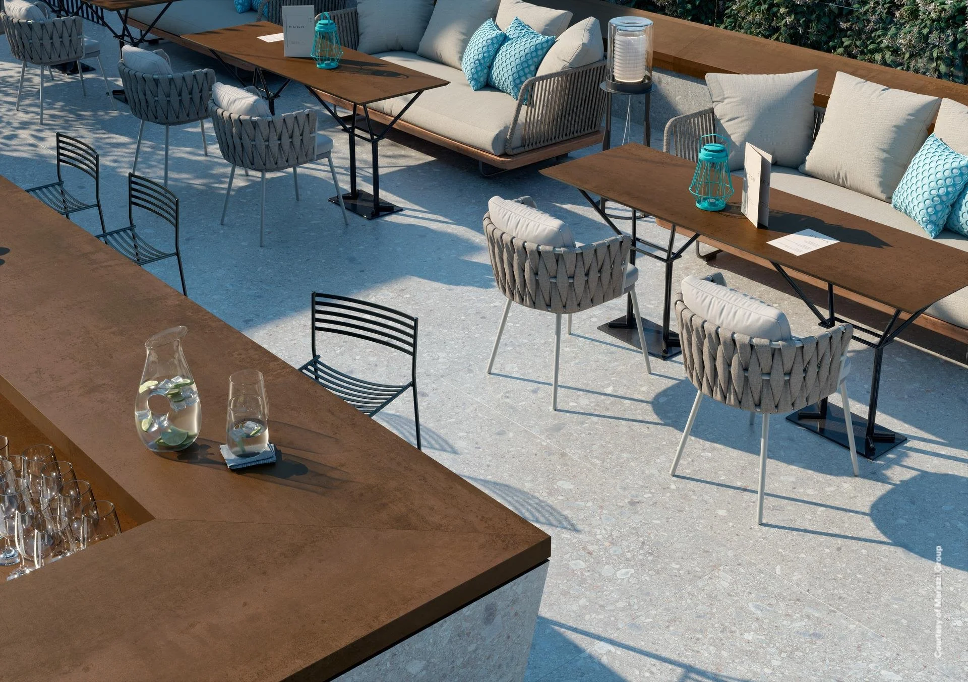 An outdoor seating area with a long wooden table and various chairs, including cushioned chairs and metal wire chairs. There is a padded sofa with decorative blue pillows along the back wall. The table has a few lanterns, menus, and a menu stand. The ground is light-colored, textured concrete, and some greenery is visible along the back.