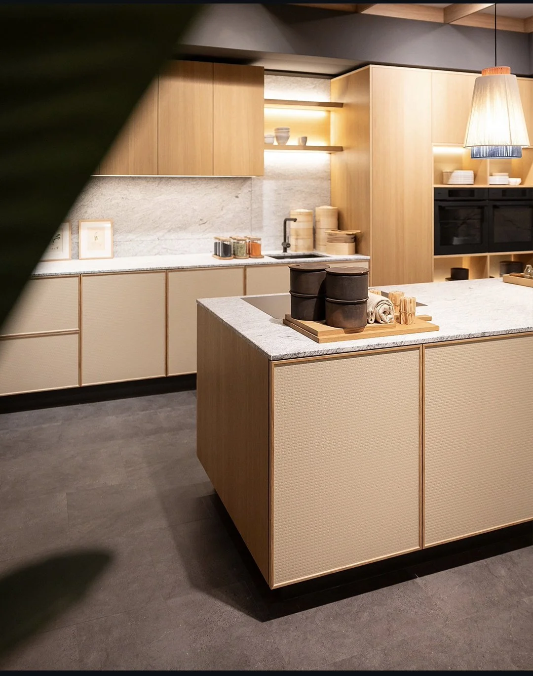 Modern kitchen with beige and wooden cabinetry, marble backsplash, and a central island with black containers and rolled towels.