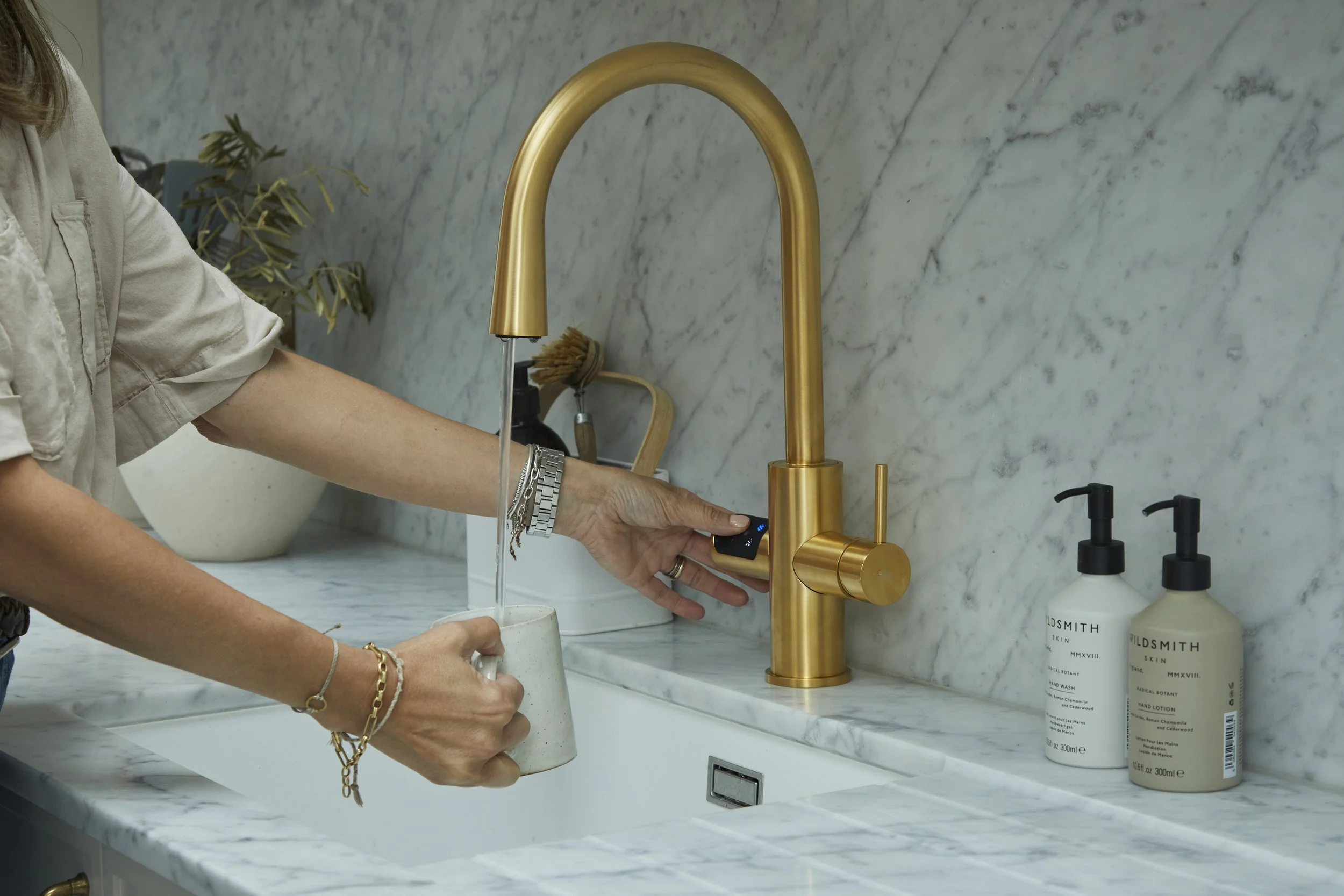 Person filling a glass with water from a gold kitchen faucet in a modern kitchen with a marble backsplash and soap bottles on the counter.