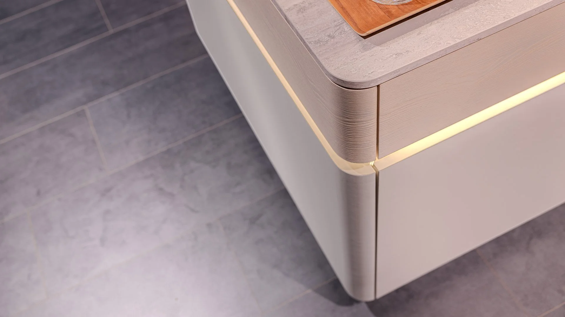 Close-up of a modern cabinet with a curved metal edge and warm LED lighting, placed on a gray tiled floor.