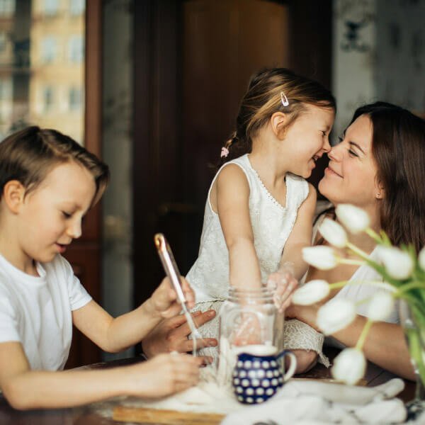 A woman and a young girl smiling and touching noses, a boy with a tablet at a table with a jar, a mug, and flowers in the foreground. Sachsenküchen lifestyle shot. German engineering kitchens.