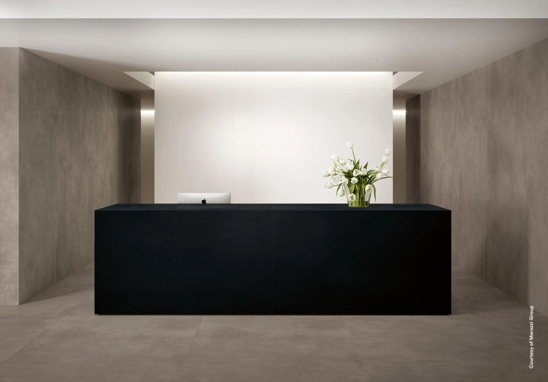 Minimalist hotel or office reception desk with a black counter, white flowers in a glass vase, and a white computer monitor against a white and beige wall.