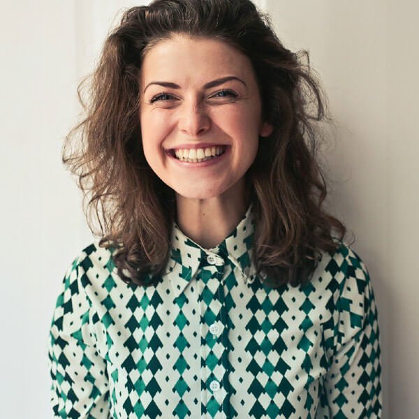 A young woman with curly brown hair smiling, wearing a patterned shirt with a white collar, standing against a light-colored wall.