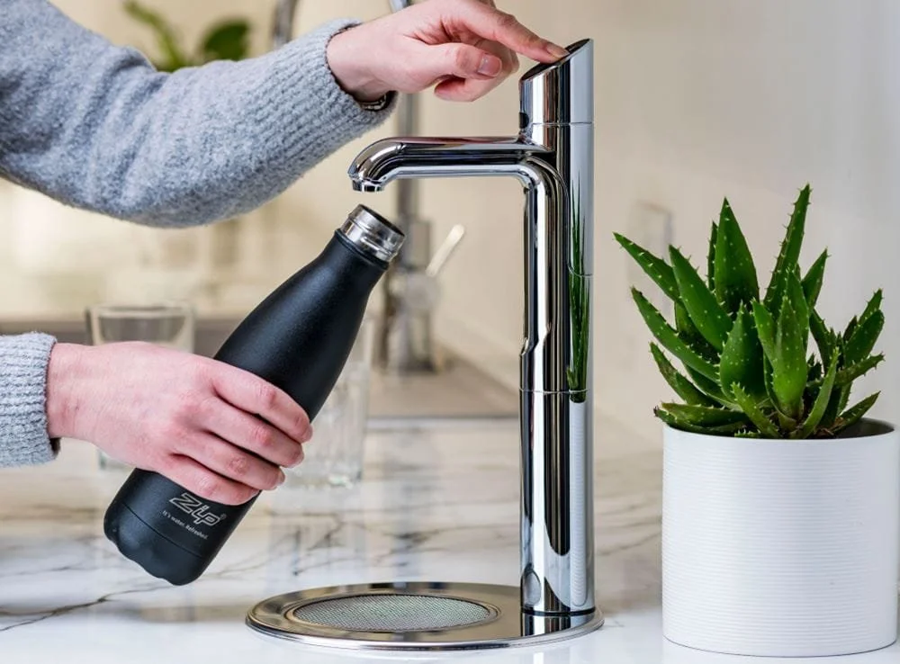 Person filling a black reusable water bottle at a modern kitchen sink with a potted aloe vera plant nearby. Zip HydroTap. Premium hot water system.
