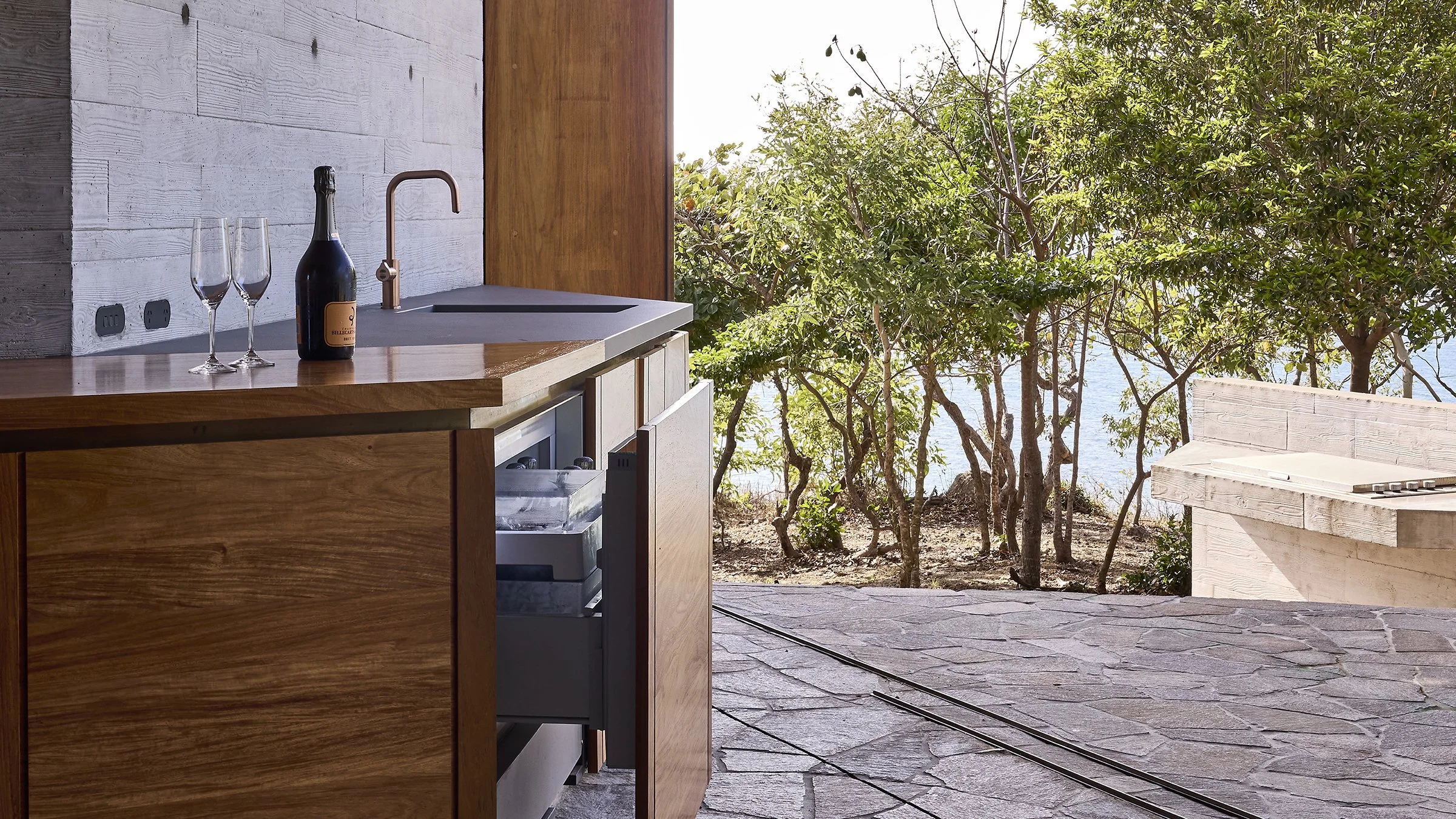 Outdoor kitchen with a wooden counter, two champagne glasses, and a bottle of champagne, with trees and water in the background.